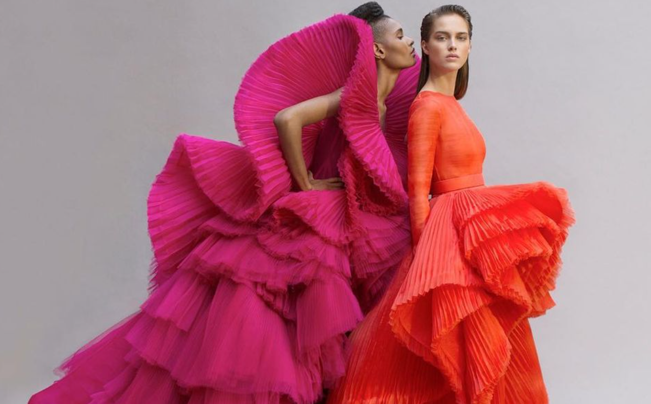 Model in a pink accordion pleated gown psoing next to another model in an orange accordion pleated gown
