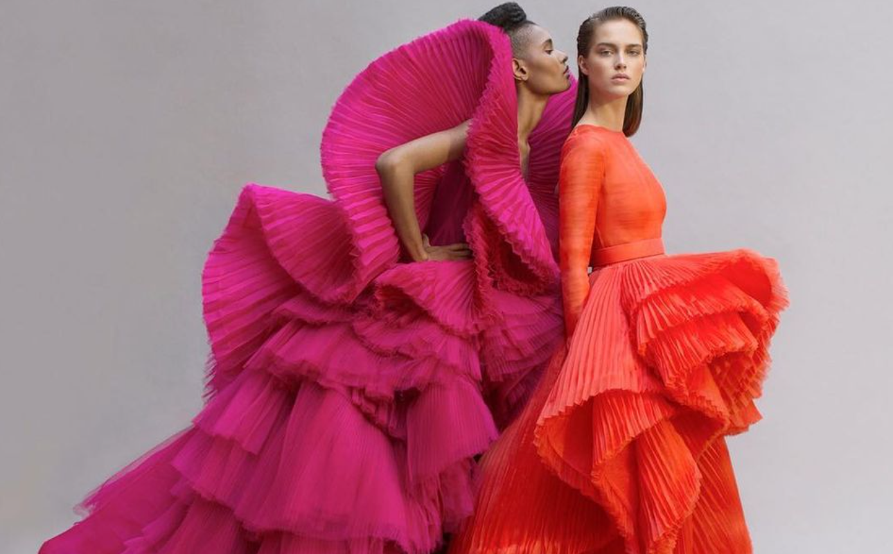 Model in a pink accordion pleated gown psoing next to another model in an orange accordion pleated gown 
